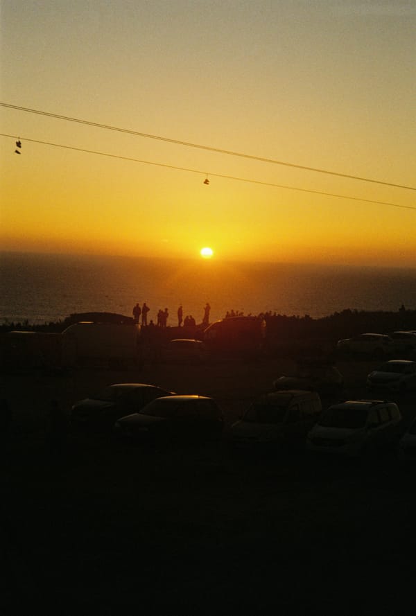People stand silhouetted against an orange sunset over the sea above a parking lot, shoes hanging from wires, Morocco