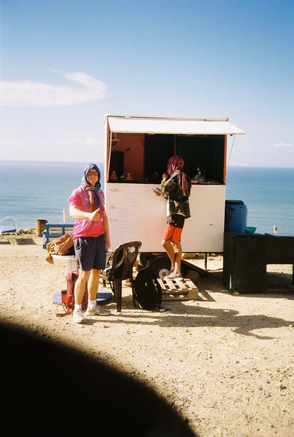Two travelers in head scarves stand by a small white kiosk on a sunny clifftop overlooking the blue sea, Morocco