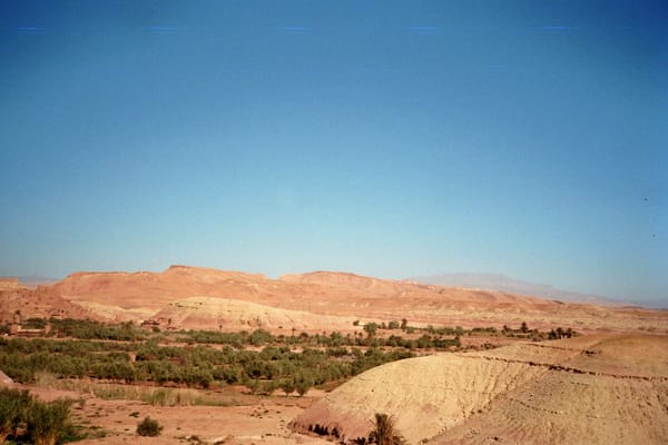 Green palm groves line a dry river valley beneath sandy hills and a faint mountain range under clear sky, Morocco