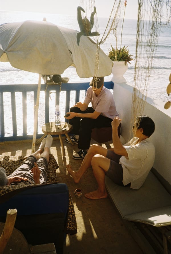 Young men relax on a sunny seaside balcony under a parasol with a sparkling ocean in the background, Morocco