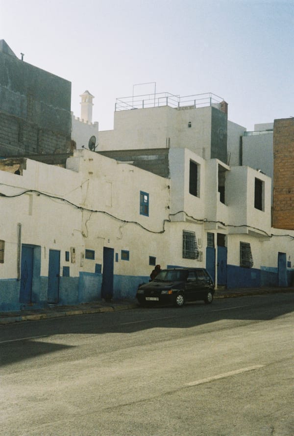 A dark car is parked on a quiet street in front of white houses with blue-painted doors and lower walls, Morocco