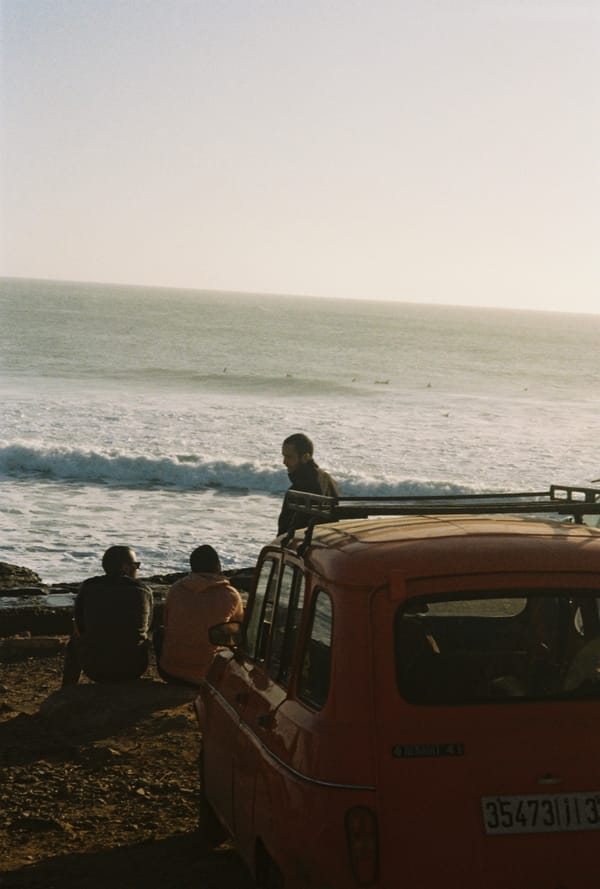 Friends sit on a rocky shore beside an old orange car watching surfers and waves in the evening light, Morocco