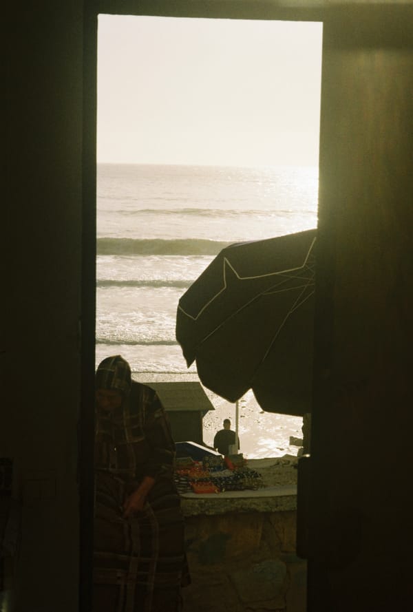 A dark doorway frames the bright ocean, a parasol and a vendor's stall where a figure in a hooded robe stands, Morocco