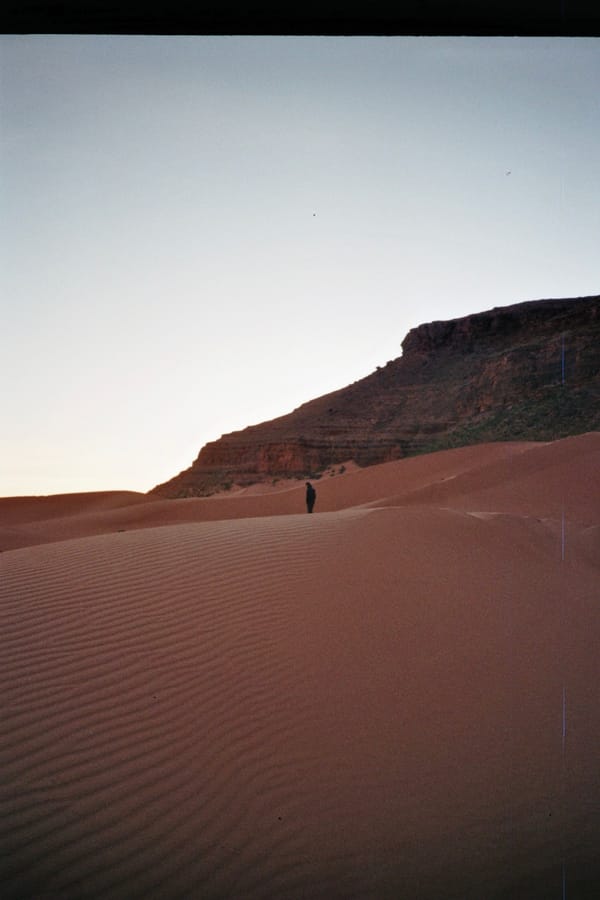 A lone figure stands on rippled red sand dunes beneath a rocky escarpment in the pale light of dusk, Morocco