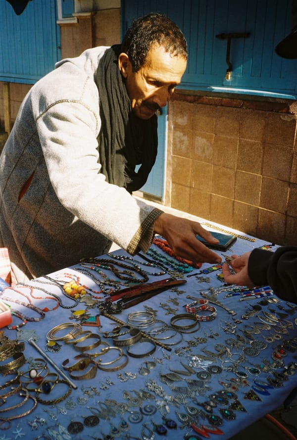 A man in a grey robe lays out silver bracelets and beaded necklaces on a blue cloth at a sunlit street stall, Morocco