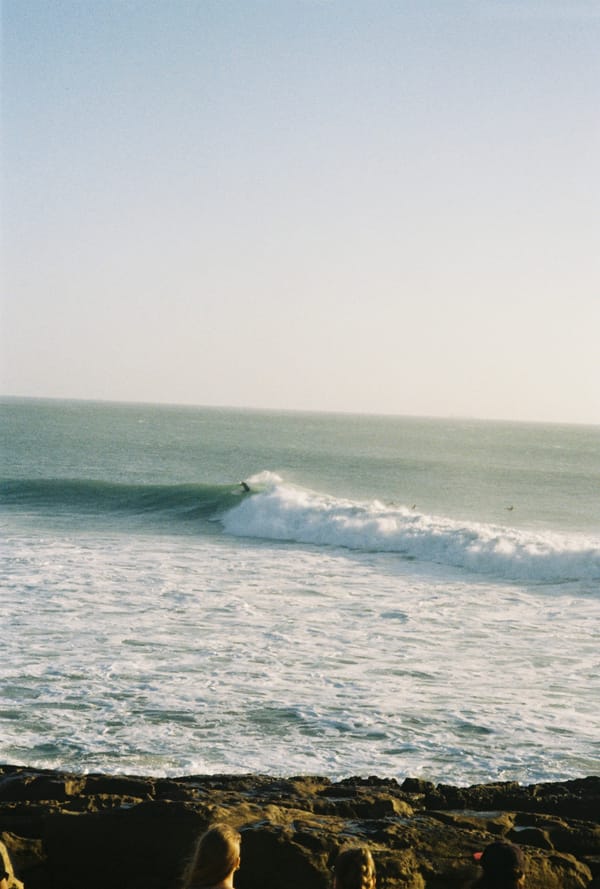 A surfer rides a long breaking wave while onlookers watch from dark rocks in the foreground in hazy light, Morocco