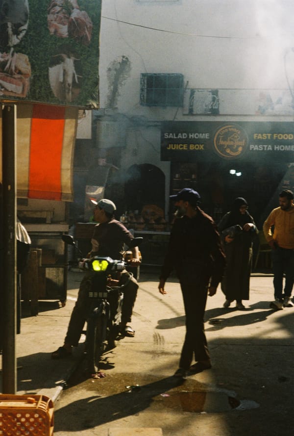 A man on a motorbike and pedestrians move through a smoky, sunlit street in front of a fast food shop, Morocco