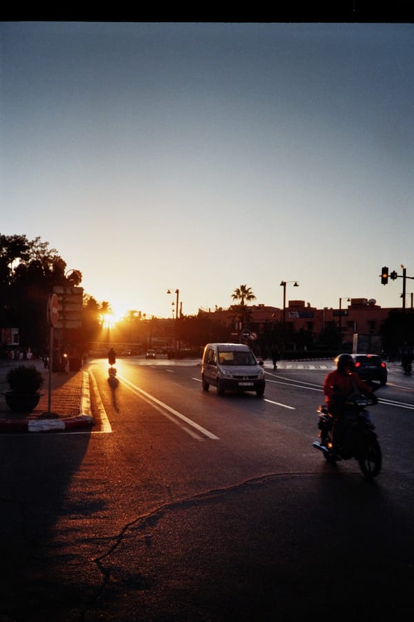 A low sun flares down a wide city road as a van and a motorcyclist drive through the warm evening glow, Morocco