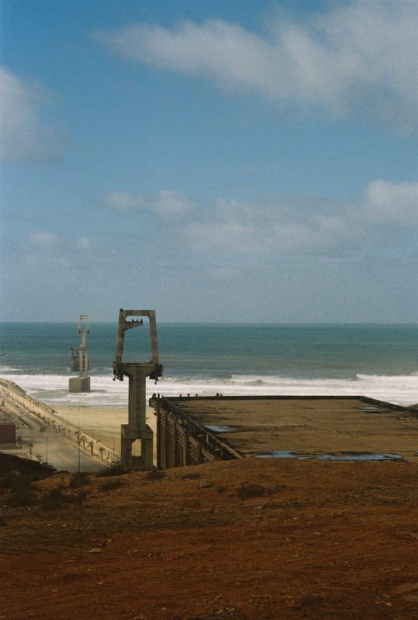 Concrete pylons and a flat-roofed structure stand on red earth above a long beach and promenade under blue sky, Morocco