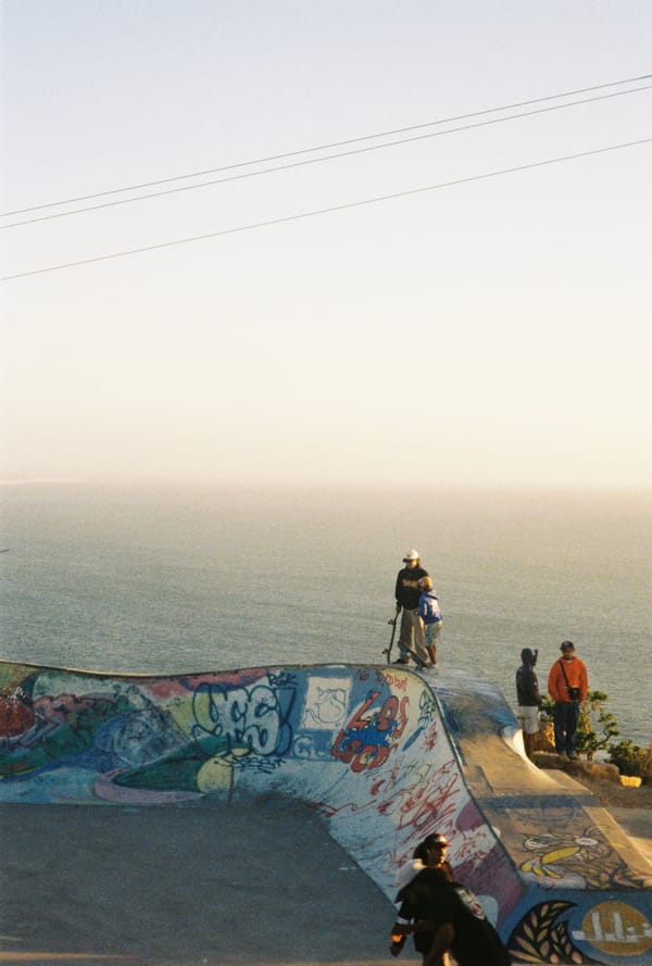 Skaters gather on the rim of a graffiti-covered concrete bowl overlooking a hazy ocean in golden light, Morocco