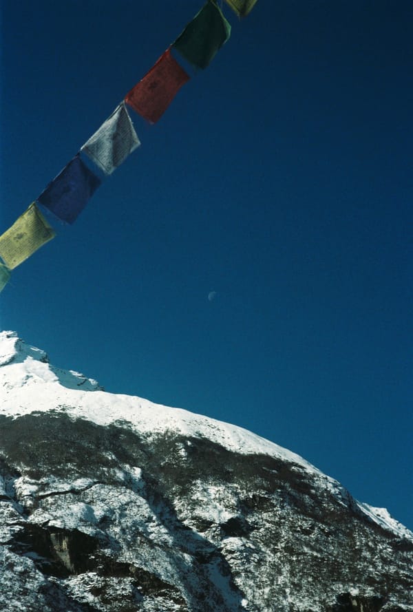 Faded prayer flags crossing a deep blue sky with a daytime moon above a snowy ridge in Nepal