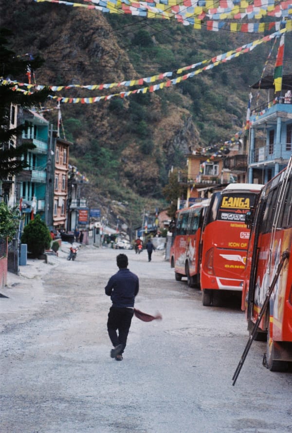 Man running down a dusty street lined with red buses under strings of prayer flags in a mountain town in Nepal