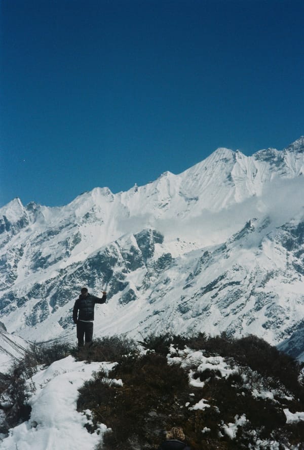 Hiker raising an arm on a snowy hilltop before a wall of jagged white peaks under clear blue sky in Nepal