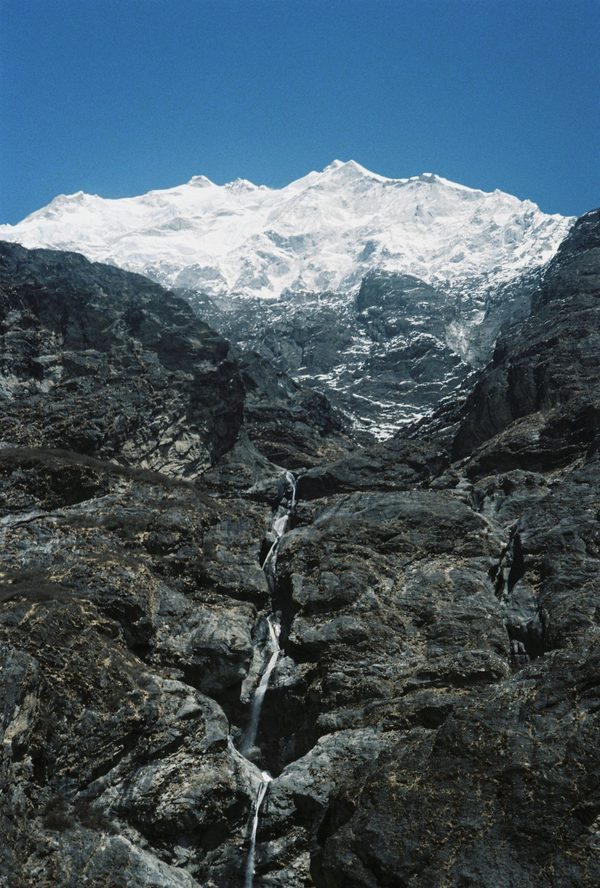Thin waterfall running down dark rock walls below a massive snow-covered summit under a deep blue sky in Nepal
