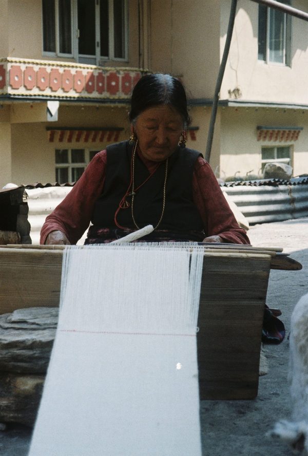 Elderly woman with gold earrings weaving a long white textile on a wooden loom in a sunny courtyard in Nepal