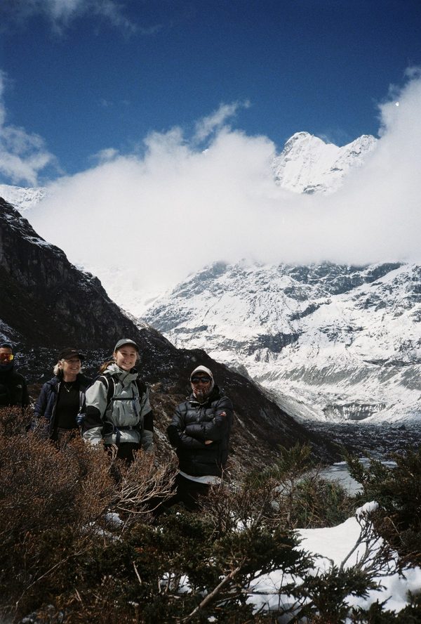 Four trekkers in warm jackets smiling among shrubs before snowy mountains wrapped in cloud in Nepal