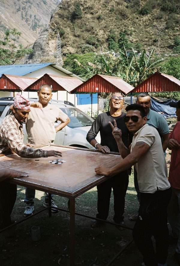 Men laughing and posing around a carrom board on a metal table outside red-roofed shelters in Nepal