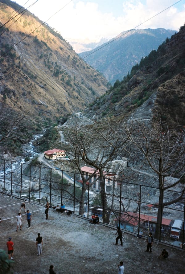 Young people playing volleyball on a fenced gravel court above a river valley between steep mountains in Nepal