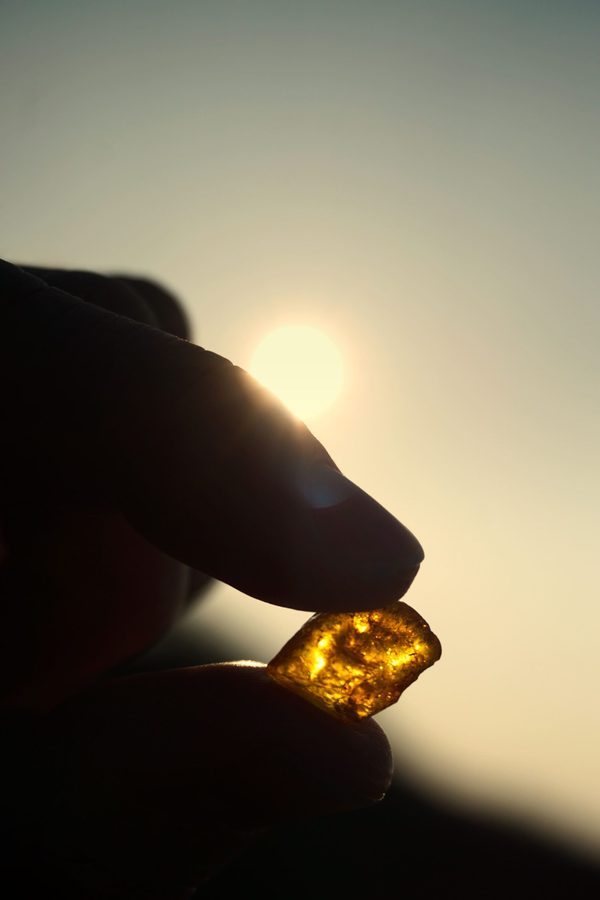 Fingers holding a small translucent golden stone up against the low sun, backlit and glowing in warm haze