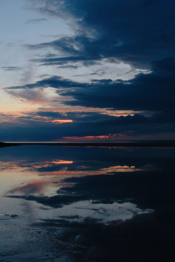 Dark blue clouds and streaks of sunset light mirrored in still shallow water on a wide, empty beach at dusk