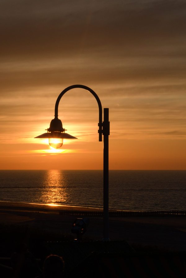 Curved promenade street lamp silhouetted against an orange sunset, the sun glowing inside its shade over the sea