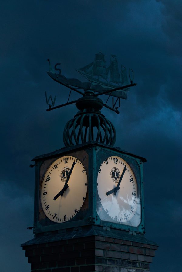 Illuminated four-sided clock tower topped by a sailing-ship weather vane against a dark, stormy blue sky
