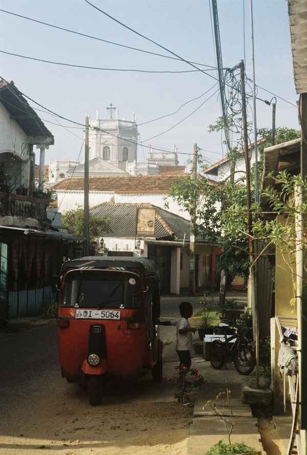 A child stands beside a red tuk-tuk on a quiet street under tangled wires, a white church tower behind, in Sri Lanka