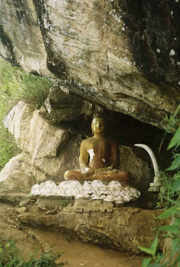 A weathered seated Buddha statue sits sheltered beneath a large overhanging rock with white lotus carvings, Sri Lanka