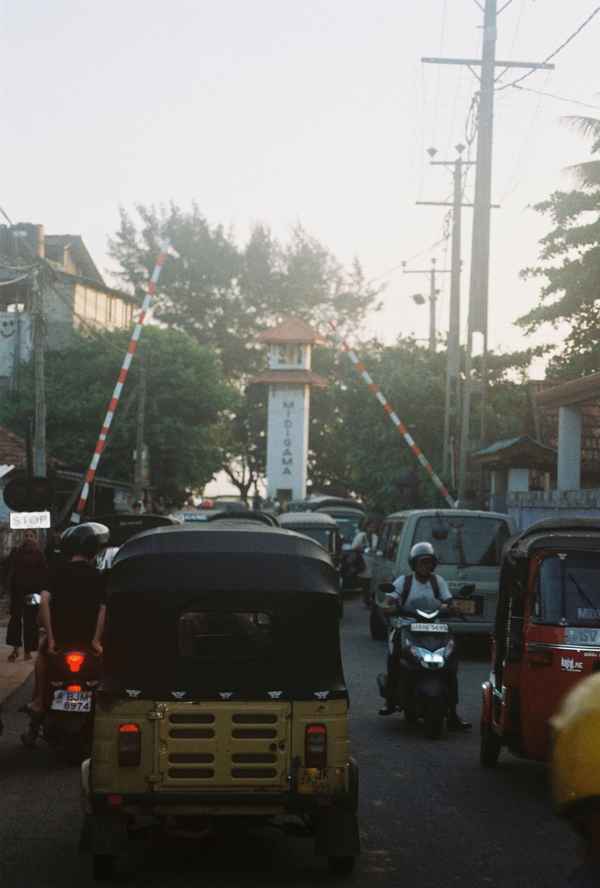 Tuk-tuks and scooters queue at a raised railway crossing barrier beside a small white tower in hazy light, Sri Lanka